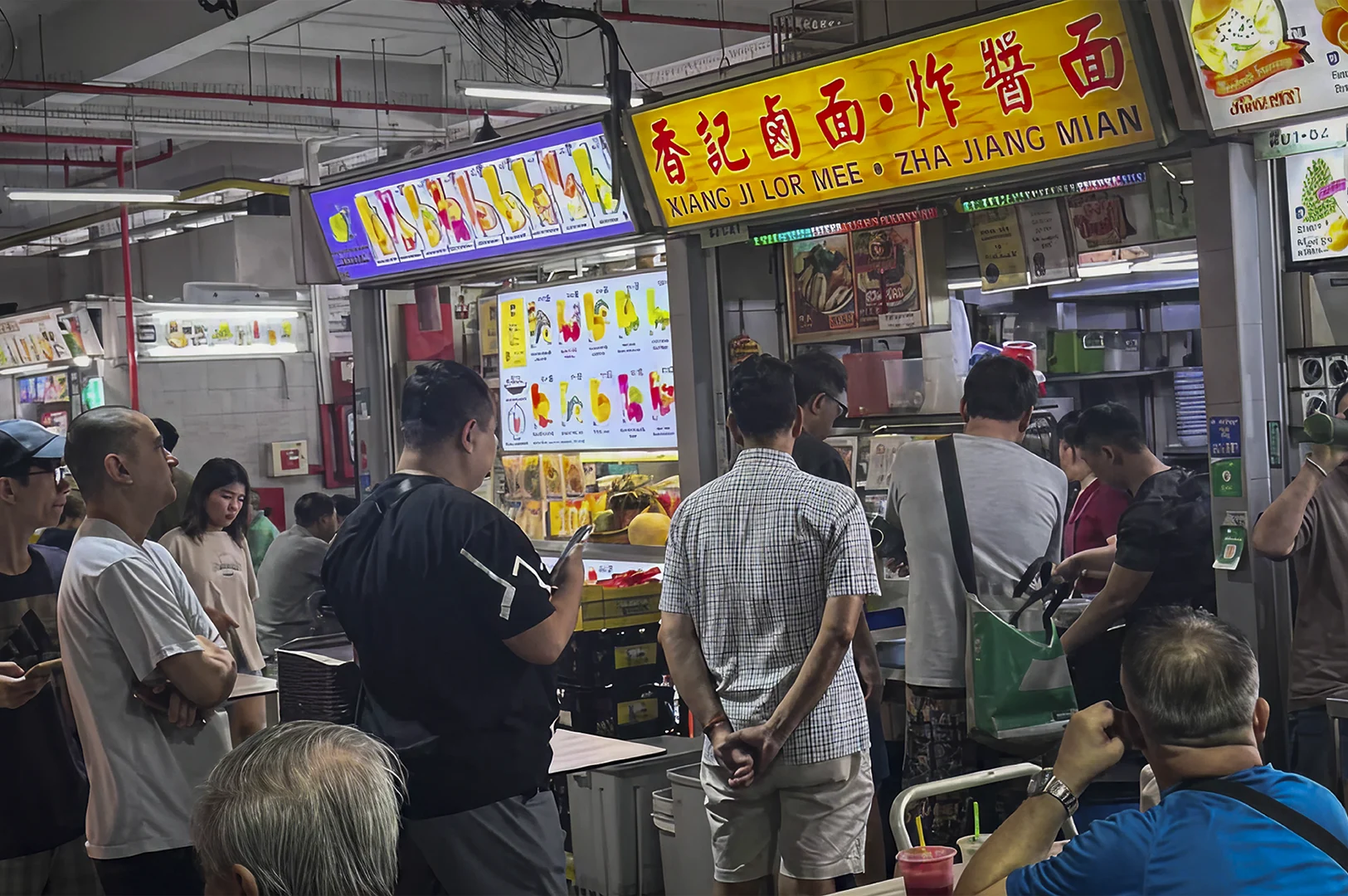 A busy, brightly lit indoor hawker center in Singapore. A line of customers stands in front of a food stall titled "XIANG JI LOR MEE • ZHA JIANG MIAN" in bold red and English lettering. Above the stall, a backlit menu displays various colorful fruit juices. People in casual summer attire wait patiently, while a man in a blue shirt is partially visible in the foreground. The atmosphere is bustling and utilitarian, with exposed ceiling pipes and fluorescent lighting.