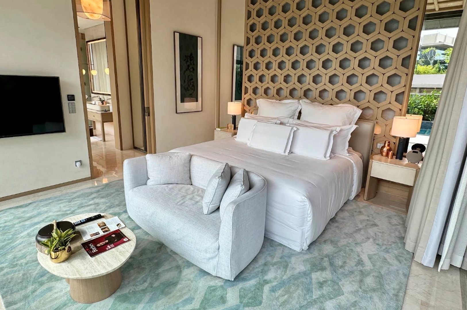 A luxurious, sunlit hotel suite featuring a plush king-sized bed dressed in crisp white linens and an abundance of pillows. The bed is backed by a striking, full-height decorative headboard with a geometric honeycomb lattice pattern in light wood. At the foot of the bed sits a modern, curved grey loveseat, facing a small round marble coffee table topped with magazines and a potted plant. The room is grounded by a textured teal and grey rug, while an open doorway in the background reveals a glimpse of a sophisticated marble bathroom.