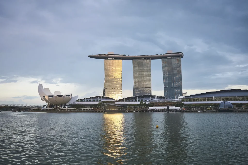 A wide landscape shot of the Marina Bay Sands resort and the ArtScience Museum in Singapore across the water at dusk. The three iconic hotel towers are topped by the cantilevered Sands SkyPark. The setting sun reflects a warm gold glow off the glass facade of the leftmost tower. In the foreground, the calm water of the bay features a single yellow buoy, while the ArtScience Museum sits to the left, resembling an open white lotus flower.