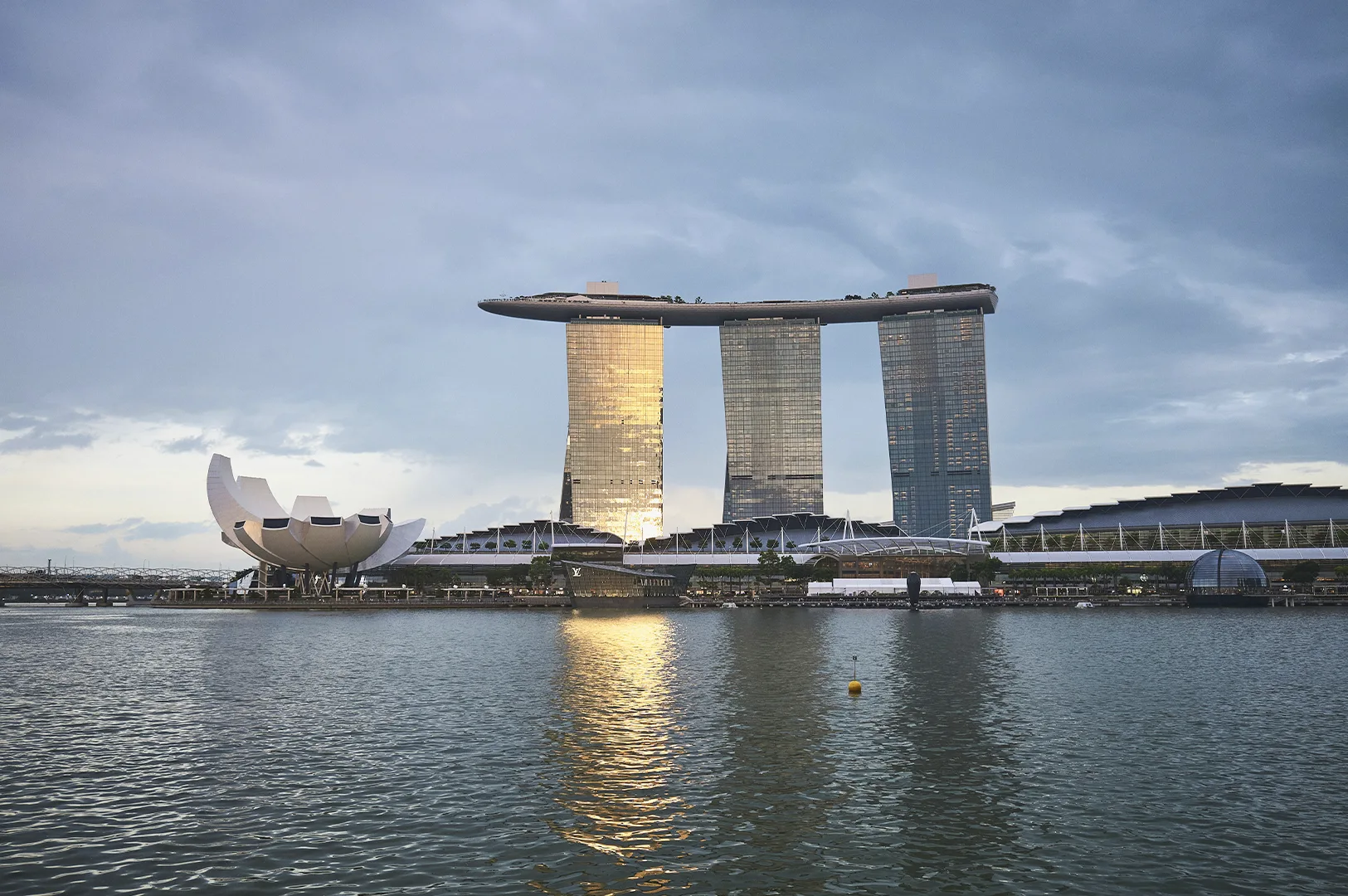 A wide landscape shot of the Marina Bay Sands resort and the ArtScience Museum in Singapore across the water at dusk. The three iconic hotel towers are topped by the cantilevered Sands SkyPark. The setting sun reflects a warm gold glow off the glass facade of the leftmost tower. In the foreground, the calm water of the bay features a single yellow buoy, while the ArtScience Museum sits to the left, resembling an open white lotus flower.