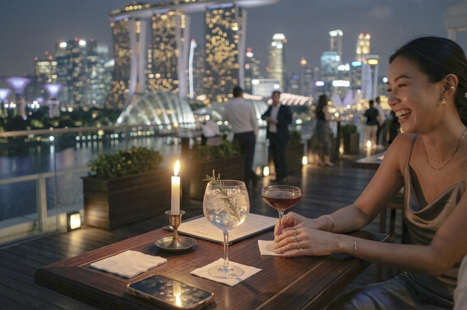 A candid, lifestyle-style photograph captures an intimate evening at a rooftop bar in Singapore, overlooking the iconic Marina Bay Sands and city skyline. In the immediate foreground, a dark wood table is set with two elegant cocktails—a gin and tonic garnished with rosemary and a dark espresso martini—alongside a flickering white candle and a smartphone reflecting the overhead lights. A woman is partially visible on the right, captured mid-laugh during a conversation, her hands resting near her drink, embodying a "quiet luxury" aesthetic in a silk slip dress and delicate gold jewelry. The background is a soft-focus bokeh of glowing skyscrapers and the Supertree Grove, with a few well-dressed patrons scattered across the wooden deck, creating a sophisticated, curated atmosphere. The lighting is a warm, atmospheric blend of candlelight and cool blue twilight, giving the scene a cinematic, influencer-inspired quality.