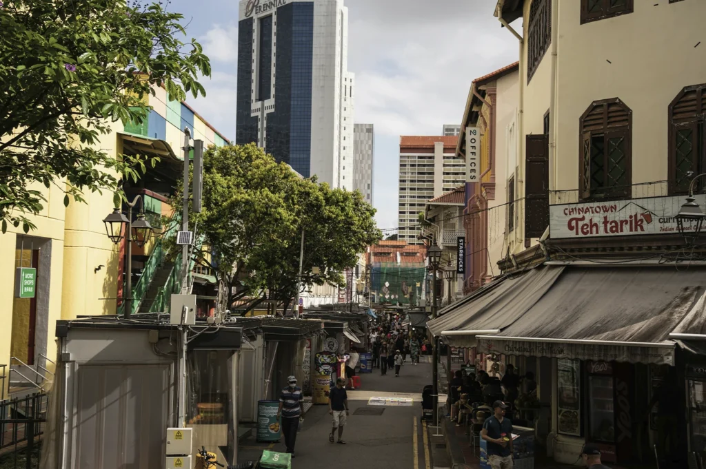 A street-level view of a bustling pedestrian lane in Singapore's Chinatown during a bright, slightly overcast day. In the foreground, the "Chinatown Teh Tarik" shop with a prominent white and red sign stands on the right, while modern high-rise buildings, including the blue-glass Perennial building, tower in the background. The narrow street is lined with market stalls under grey awnings, and a few people are seen walking through the area, blending the traditional low-rise shophouse architecture with the city's sleek urban skyline.