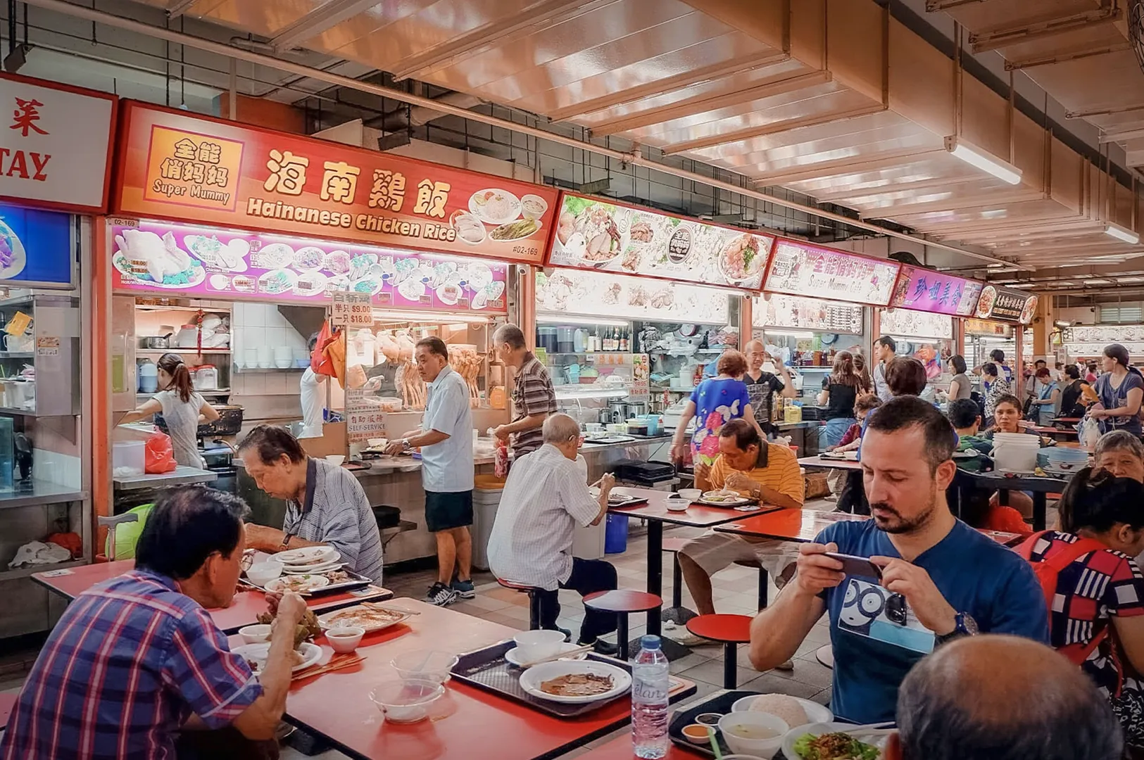 A bustling scene at a Singapore hawker centre featuring a Hainanese Chicken Rice stall, with diners seated at red tables enjoying traditional local dishes in a vibrant, high-traffic food court.