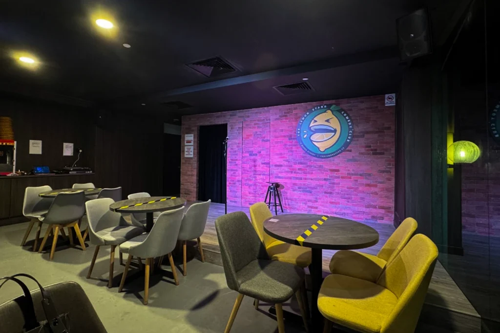 The interior of "The Lemon Stand Comedy Club" before a show. The stage has a rustic red-brick backdrop featuring a large circular logo of a laughing lemon. A single microphone stand and a tall black stool are centered on the wooden platform. The seating area consists of modern grey and yellow chairs arranged around small dark wood tables, some of which have yellow and black hazard tape across them.