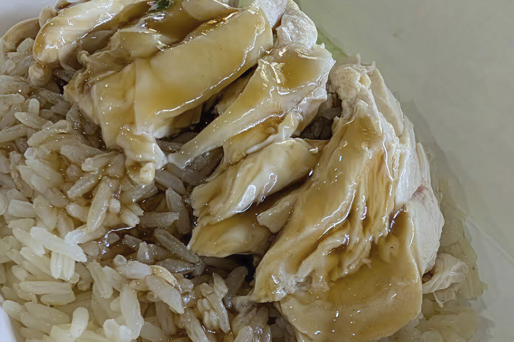 A close-up, top-down view of Hainan Chicken Rice. The dish consists of a bed of seasoned, oily rice topped with succulent, sliced poached chicken. The chicken skin is glistening with a light soy-based sauce and sesame oil glaze, emphasizing its tender texture.