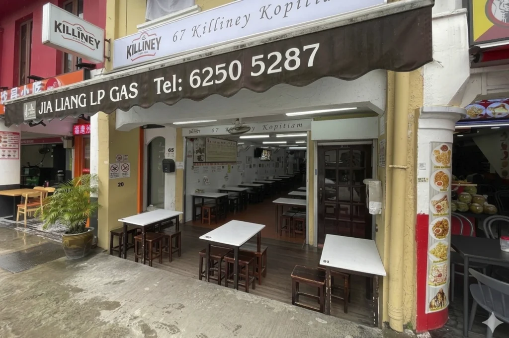 The storefront of Killiney Kopitiam at 67 Killiney Road. The traditional Singaporean coffee shop features a white awning with dark brown scalloped edges, displaying the shop's name and phone number. Below the awning, several simple white rectangular tables with dark wooden stools are arranged both outside on the sidewalk and inside the brightly lit, narrow interior. The building has a pale yellow facade with a large pillar on the right side decorated with colorful food photography posters. The ground is a grey concrete sidewalk, showing damp patches from recent rain.
