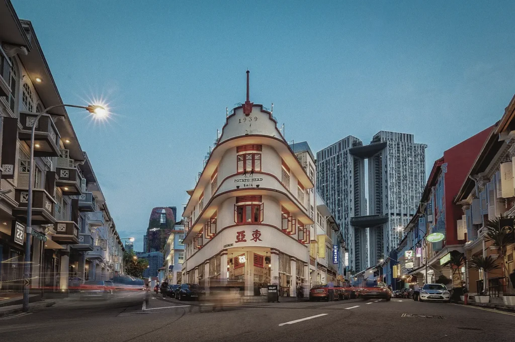 The iconic heritage building at the corner of Keong Saik Road in Singapore. The white, wedge-shaped Art Deco building features red window frames and the year "1939" inscribed at the top. The street level is vibrant with warm lights from the "Potato Head" restaurant. In the background, the modern, towering Pinnacle@Duxton skyscrapers create a sharp contrast between historic architecture and contemporary urban design under a deep blue twilight sky.