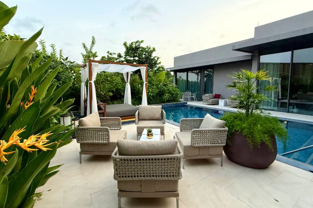 An expansive private outdoor terrace at dusk, featuring a sparkling blue swimming pool that wraps around a contemporary stone patio. In the foreground, four stylish woven-back armchairs are arranged around a small white side table, while a wooden cabana with white billowing curtains and a large daybed offers a secluded lounging spot in the background. Lush tropical greenery, including vibrant orange bird-of-paradise flowers and dense palms, encloses the space, providing privacy against the backdrop of a modern, glass-walled villa.