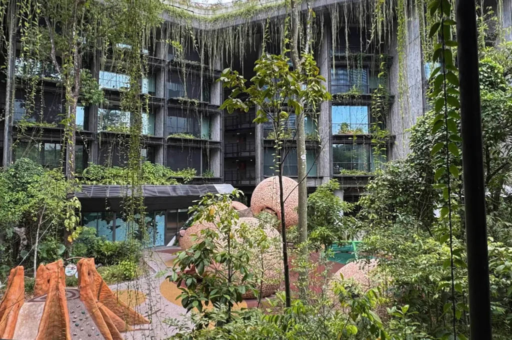 A multi-story modern building features a facade covered in vertical greenery and hanging vines. In the central courtyard, large, rounded, textured sculptures resembling giant seeds or stones sit among tropical trees and a playground area with orange, jagged climbing structures.