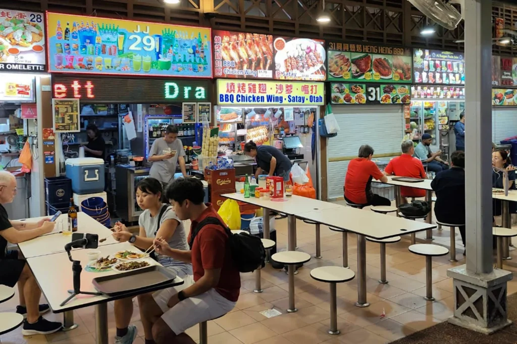 A vibrant evening scene inside a Singaporean hawker center, showing several brightly lit food stalls with colorful illuminated menus. Stalls featured include "JJ 129" for drinks and "Heng Heng BBQ" specializing in BBQ chicken wings and satay. In the foreground, customers are seated at long, simple white tables on round stools; one man on the left checks his phone while a couple nearby eats with chopsticks, capturing the casual, communal, and energetic atmosphere of local night dining.
