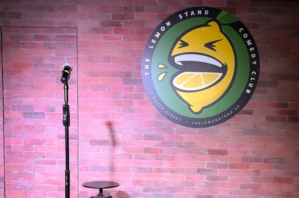 The stand-up comedy stage at The Lemon Stand Comedy Club in Singapore, featuring a microphone on a stand, a small stool, and the club's circular laughing lemon logo mounted on a red brick wall.