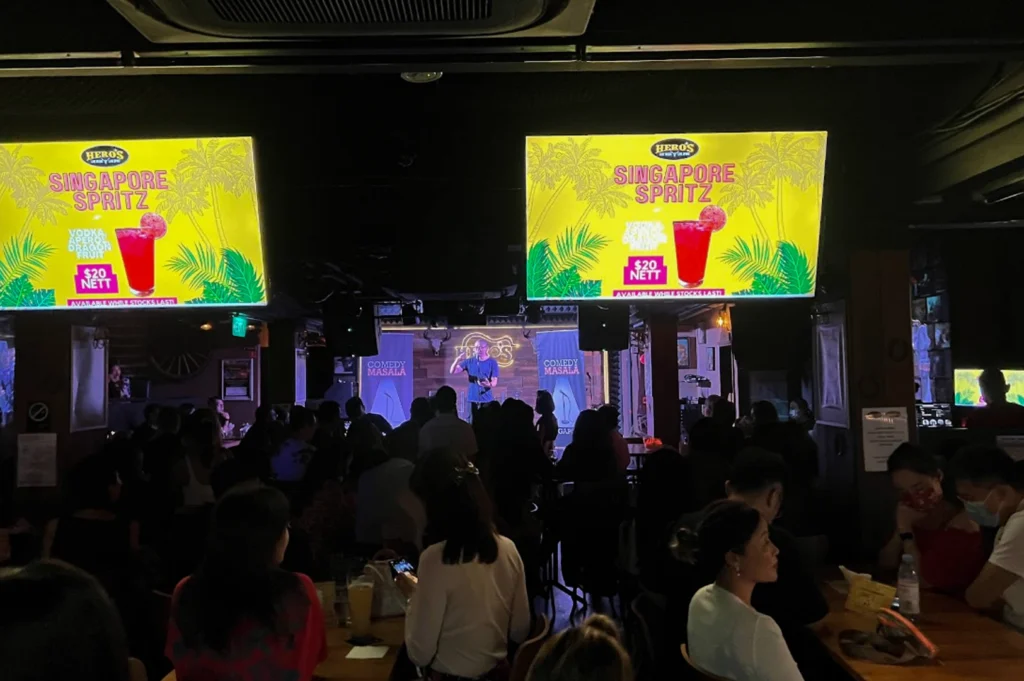 A crowded, dark bar during a performance at "Comedy Masala." Two large glowing television screens hang from the ceiling, displaying a bright yellow advertisement for a "$20 Nett Singapore Spritz." In the distance, a comedian stands on a small stage between two blue banners. The room is filled with patrons seated at wooden tables, their silhouettes visible against the stage light and the glow of the overhead monitors.