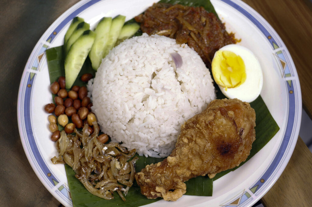 A traditional presentation of Nasi Lemak served on a white plate with a green banana leaf liner. In the center is a mound of coconut milk rice, surrounded by classic sides: a crispy fried chicken drumstick, a hard-boiled egg half, sliced fresh cucumbers, a pile of fried anchovies (ikan bilis) with peanuts, and a dollop of deep red spicy sambal.