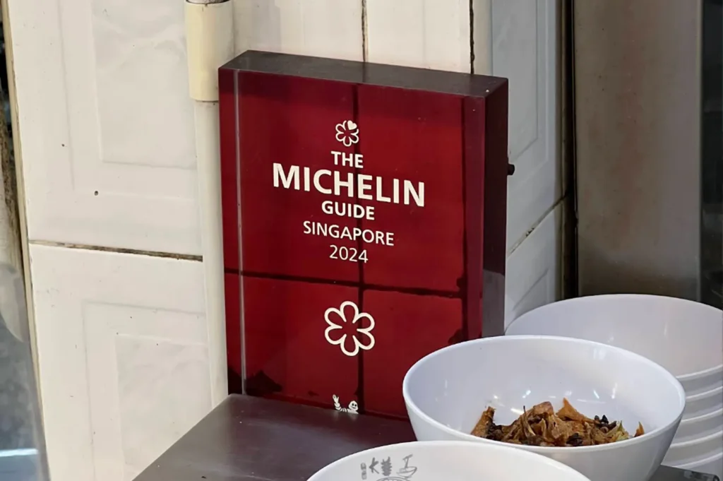 Transparent acrylic Michelin Guide plaque for Singapore 2024. The plaque features the iconic white Michelin Star symbol and text. It is placed on a stainless steel counter next to a stack of clean white ceramic bowls, one of which contains fried garnishes. The setting suggests a high-quality, recognized food stall or casual eatery.