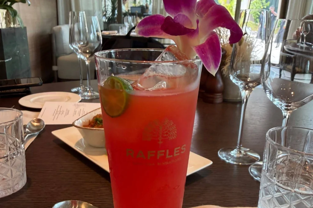 A close-up, vibrant shot of a tall glass filled with a bright pink tropical cocktail, likely a Singapore Sling, condensation beading on the outside. The glass features an etched "Raffles" logo and is garnished with a fresh green lime slice and a dramatic purple orchid perched on top. The drink sits on a dark wooden restaurant table surrounded by elegant crystal glassware, polished silverware, and a small white dish of food, with the blurred interior of a high-end dining room visible in the background.