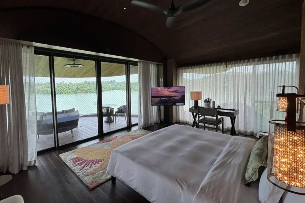 The interior of a luxury "seed pod" villa features dark wood floors and a curved wooden ceiling. A large king-sized bed with white linens faces floor-to-ceiling glass sliding doors that open onto a private wooden deck. The deck overlooks a wide lake and forest. A modern desk, a flat-screen TV on a swivel stand, and an ornate, glowing floor lamp complete the room.