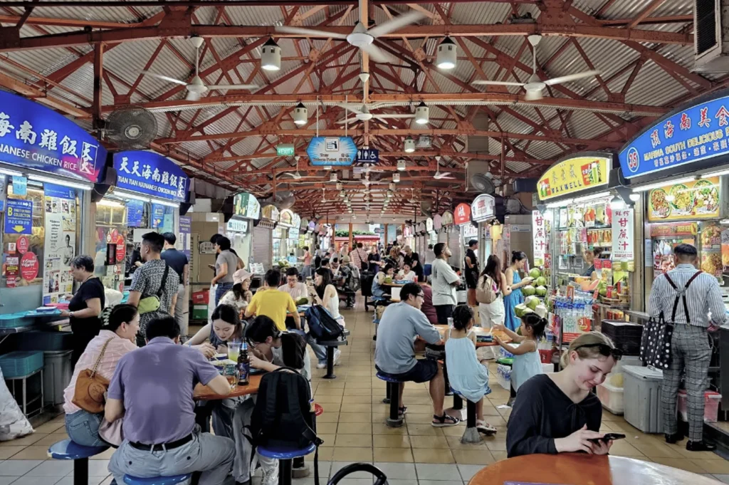 An interior wide shot of the famous Maxwell Food Centre in Singapore, showcasing its iconic red-painted steel roof trusses and rows of fluorescent-lit food stalls. The legendary "Tian Tian Hainanese Chicken Rice" stall is visible on the left. The center is filled with a diverse crowd of locals and tourists seated at orange circular tables, creating a lively, noisy, and authentic atmosphere typical of Singapore’s most popular culinary landmarks.