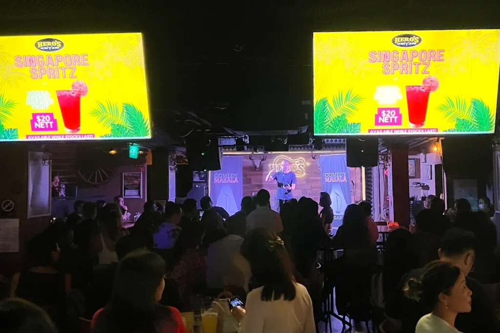 A live stand-up performance at Comedy Masala in Singapore, showing a comedian on stage at Hero's bar with a bustling audience and large digital screens displaying "Singapore Spritz" drink promotions.