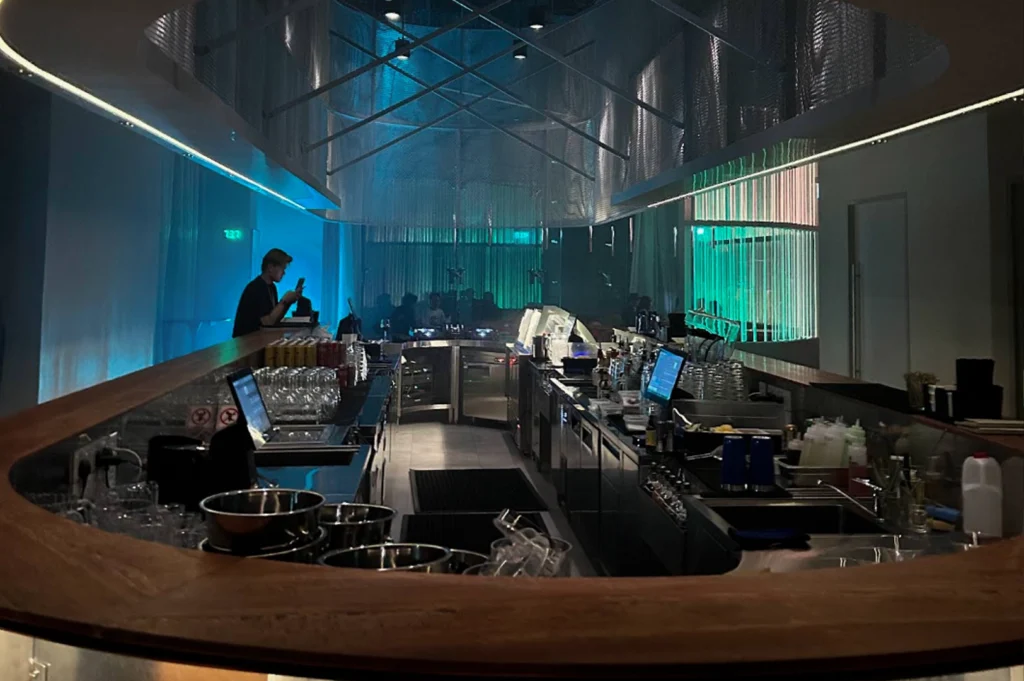 A view from behind a curved wooden bar counter looking into a modern, stylish lounge. The space is illuminated with cool blue and green neon lighting. In the background, vertical light rods create a luminous green wall. The bar area is clean and organized with stainless steel equipment, glassware, and monitors. A bartender stands to the left, silhouetted against the blue light while checking a device.