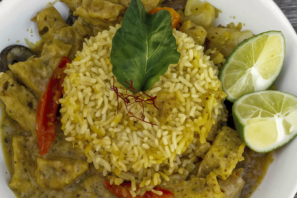 A bright and colorful dish featuring yellow turmeric rice molded into a dome shape. The rice is topped with a single green leaf and delicate red saffron threads. It is surrounded by a rich, thick curry containing pieces of tofu and vegetables, served with two fresh lime halves on the side.