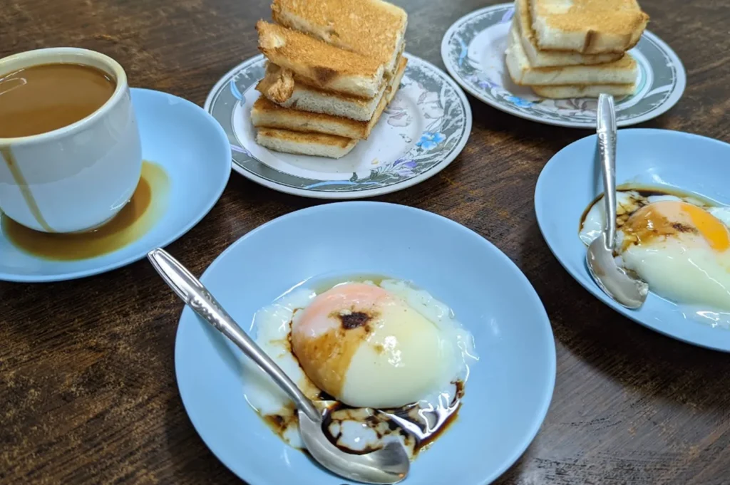A classic Nanyang breakfast spread on a dark wooden table. It includes two blue plates, each holding two soft-boiled eggs drizzled with dark soy sauce and white pepper. Two floral-patterned plates hold stacks of crispy, golden-brown kaya toast. A white ceramic mug filled with thick, milky "kopi" (coffee) sits on a matching blue saucer, with a small coffee drip on the side of the cup.