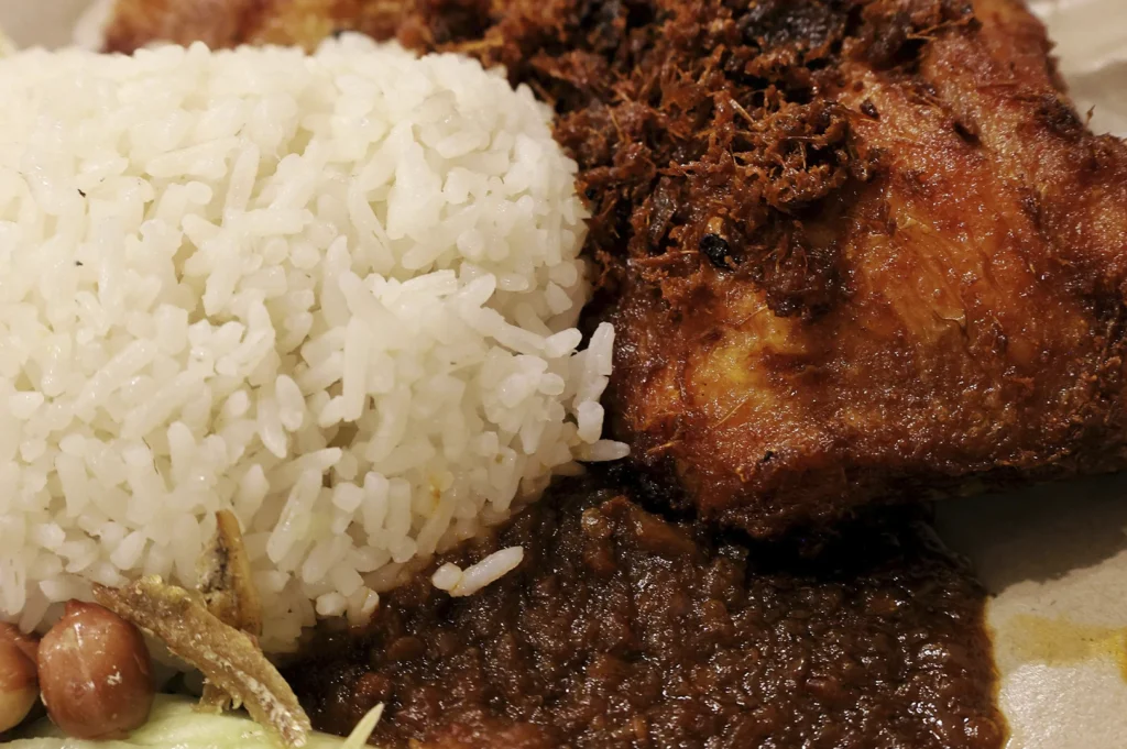 An extreme close-up of a plate of Nasi Lemak, highlighting its rich textures and colors. The frame is dominated by a portion of fluffy white coconut rice, a piece of deep-fried chicken with a thick, crispy spice crust, and a generous dollop of dark red, chunky sambal chili. In the bottom corner, traditional accompaniments like crunchy fried anchovies (ikan bilis), roasted peanuts, and a sliver of fresh cucumber are visible, emphasizing the dish's spicy and savory appeal.