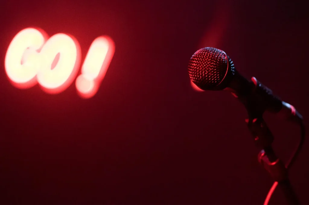 A close-up of a professional microphone on a stand in a comedy club, illuminated by a moody red light with a glowing "GO!" neon sign blurred in the background.
