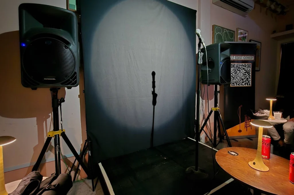 A close-up, low-angle shot of an empty stage set up for East Coast Comedy. A single spotlight hits a dark grey backdrop, casting a long shadow of a microphone stand. Two large Mackie speakers on tripod stands flank the stage. In the foreground, a wooden table holds small, modern portable lamps and cans of Coca-Cola, with a banner featuring a QR code visible in the background.