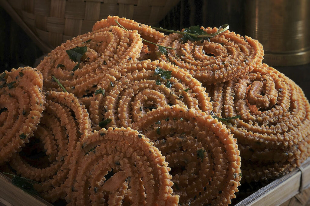 A stack of Murukku, a savory and crunchy Indian snack. The deep-fried rings have a distinct spiral shape and a rough, spiked texture. They are golden-brown and speckled with small green curry leaves and black sesame seeds, resting in a woven basket.