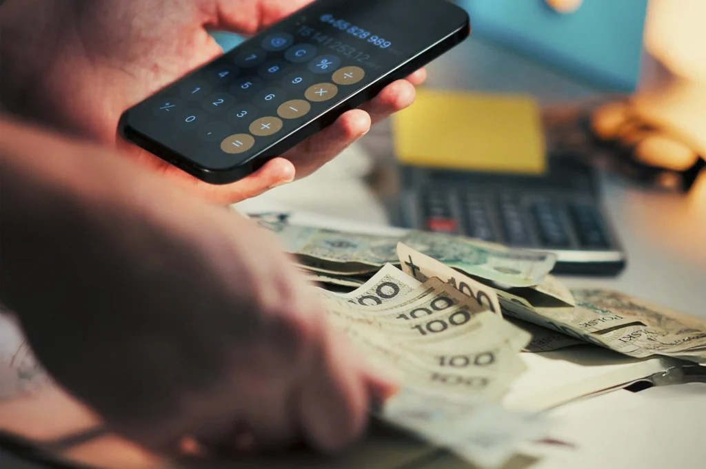 A person’s hands. One hand holds a smartphone displaying a calculator app with large numbers, while the other hand rests on a fan of Polish Złoty banknotes (100 denomination) spread across a desk. In the blurred background, a physical calculator and a yellow sticky note are visible under warm, atmospheric lighting, suggesting a small business owner or individual managing finances.