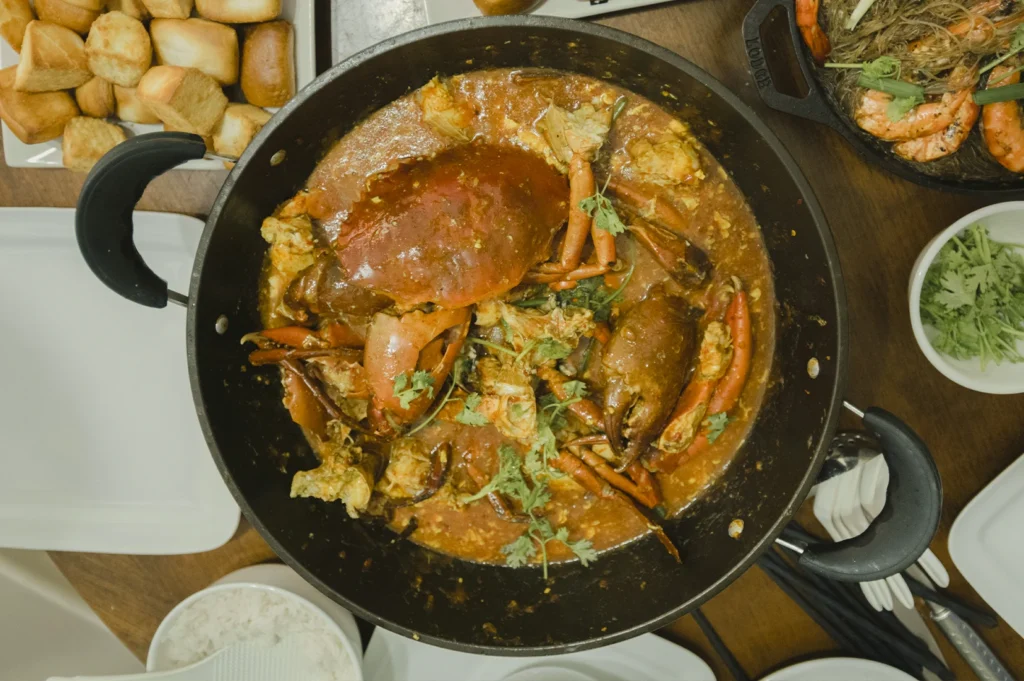 An overhead "flat lay" shot of a large black wok filled with Singaporean Chili Crab, the country's national seafood dish. Two large mud crabs are submerged in a thick, vibrant orange-red gravy flecked with egg ribbons and garnished with fresh cilantro. Surrounding the wok are side dishes including a plate of golden-brown fried mantou (buns) for dipping, a bowl of white rice, and a small dish of extra cilantro, all arranged on a wooden dining table.