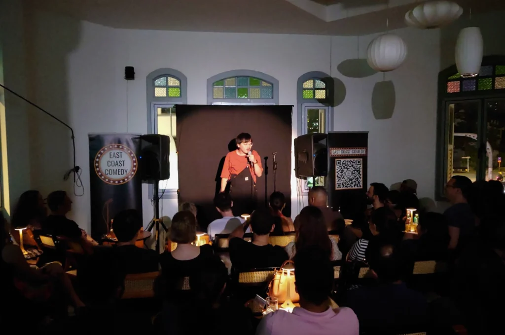 A live performance at East Coast Comedy. A young comedian in a red t-shirt and black overalls stands center stage under a bright spotlight, speaking into a handheld microphone. They are backed by a simple black curtain. To their left and right are large PA speakers on stands and banners with the "East Coast Comedy" logo and a QR code. The audience is seated in a dimly lit room with traditional patterned windows in the background.