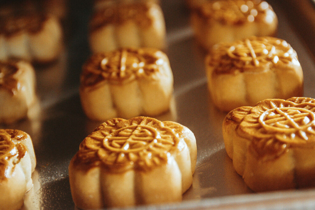 A close-up of several baked mooncakes arranged in rows on a metal baking tray. Each mooncake has a glossy, golden-brown crust with intricate traditional Chinese patterns and characters embossed on the top. The lighting is warm, highlighting the detailed floral designs on the pastry.