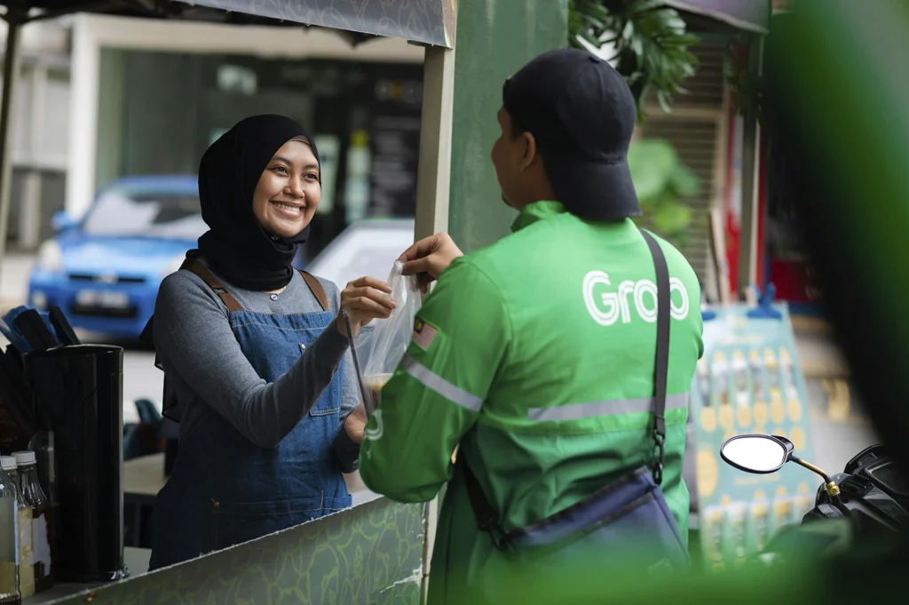 A cheerful interaction between a food vendor and a delivery rider. A woman wearing a black hijab and a denim apron smiles warmly as she hands a plastic bag containing a drink to a GrabFood delivery rider. The rider is wearing the signature bright green "Grab" jacket with reflective stripes and a black cap