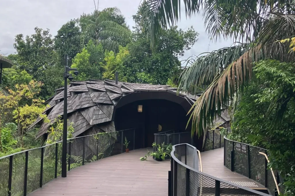 A ground-level view of a boardwalk leading to the entrance of a unique villa. The structure is covered in dark, overlapping wooden shingles, giving it a scaly, organic appearance like a pinecone or seed. Lush tropical plants and palm fronds frame the walkway, and a black lamppost stands to the left.