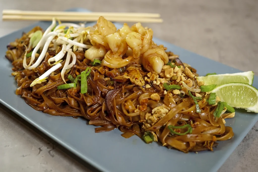 A close-up, angled shot of a plate of Char Kway Teow, a popular stir-fried noodle dish. The dark, glossy flat rice noodles are tossed with bean sprouts, chives, and bits of egg, topped with several large, juicy poached prawns and a sprinkle of crushed peanuts. Two lime wedges sit on the side of the blue rectangular plate, suggesting a balance of smoky "wok hei" flavor with a bright, acidic finish.