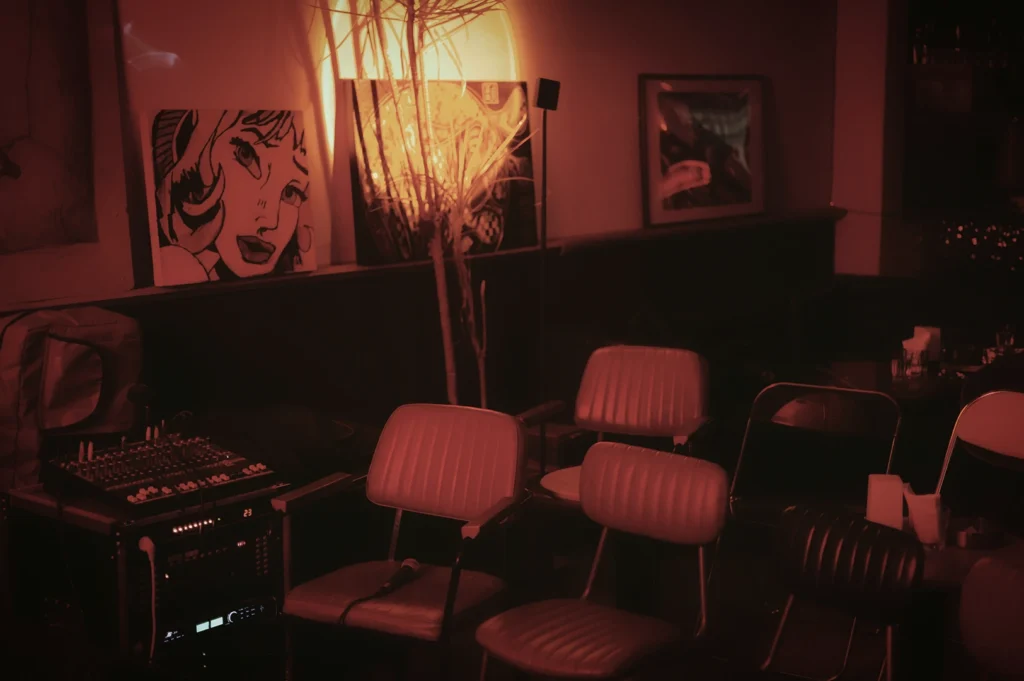 The interior of an empty comedy bar under moody red lighting, featuring rows of chairs, a sound mixer, and pop-art illustrations on the wall, creating a cozy and artistic atmosphere.