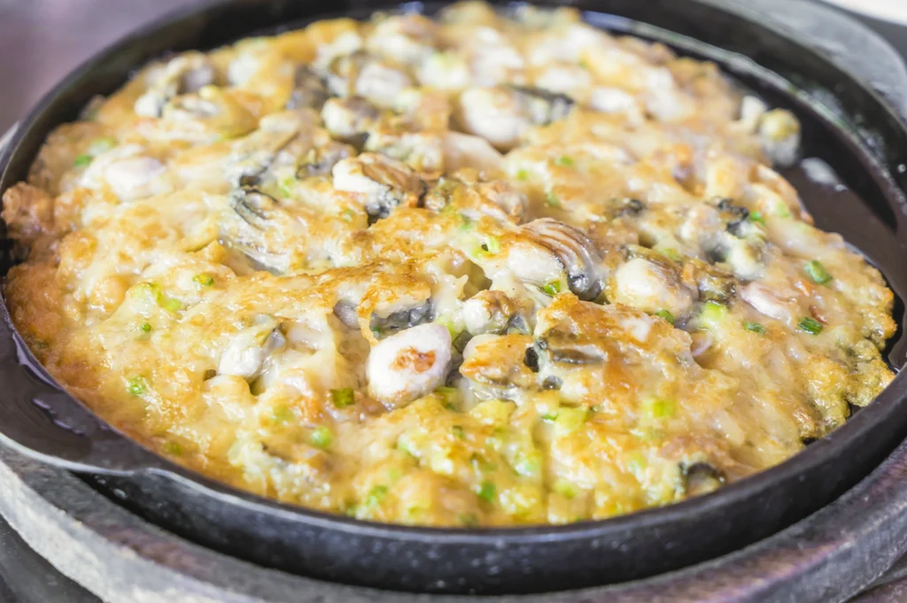 A top-down view of a sizzling Oyster Omelette (Orh Luak) served in a round black cast-iron skillet. The omelette is a textured mix of crispy fried batter and soft, gooey starch, studded with numerous plump, greyish-white oysters and garnished with chopped green spring onions. The golden-brown edges of the egg contrast with the tender centers of the oysters, capturing the classic hawker dish just as it is served hot.