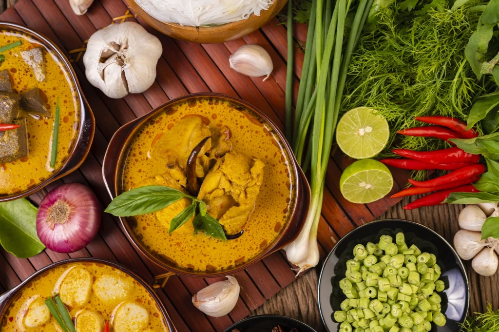 A vibrant, top-down culinary arrangement featuring a bowl of rich yellow Thai-style chicken curry as the centerpiece. The curry is topped with a fresh green basil leaf and a dried red chili, surrounded by raw ingredients such as whole garlic bulbs, a red onion, lime halves, fresh green beans, and long stalks of lemongrass. The elements are meticulously placed on a dark wooden slated mat, creating a high-contrast and appetizing display of Southeast Asian flavors.