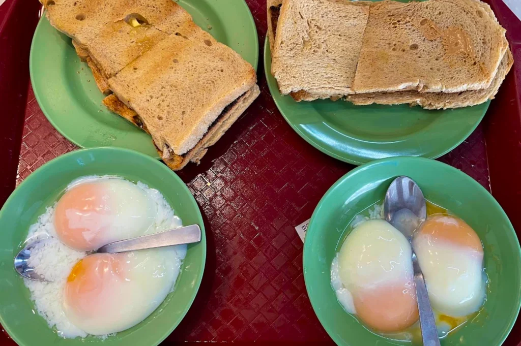 A classic Singaporean breakfast set displayed on a red plastic tray with bright green melamine plates. The set includes two plates of thick-cut, toasted brown bread slathered with kaya (coconut jam) and butter, and two small bowls containing "soft-boiled" eggs with runny yolks and whites. Stainless steel spoons rest in the eggs, ready to be seasoned with dark soy sauce and white pepper for a traditional morning meal.