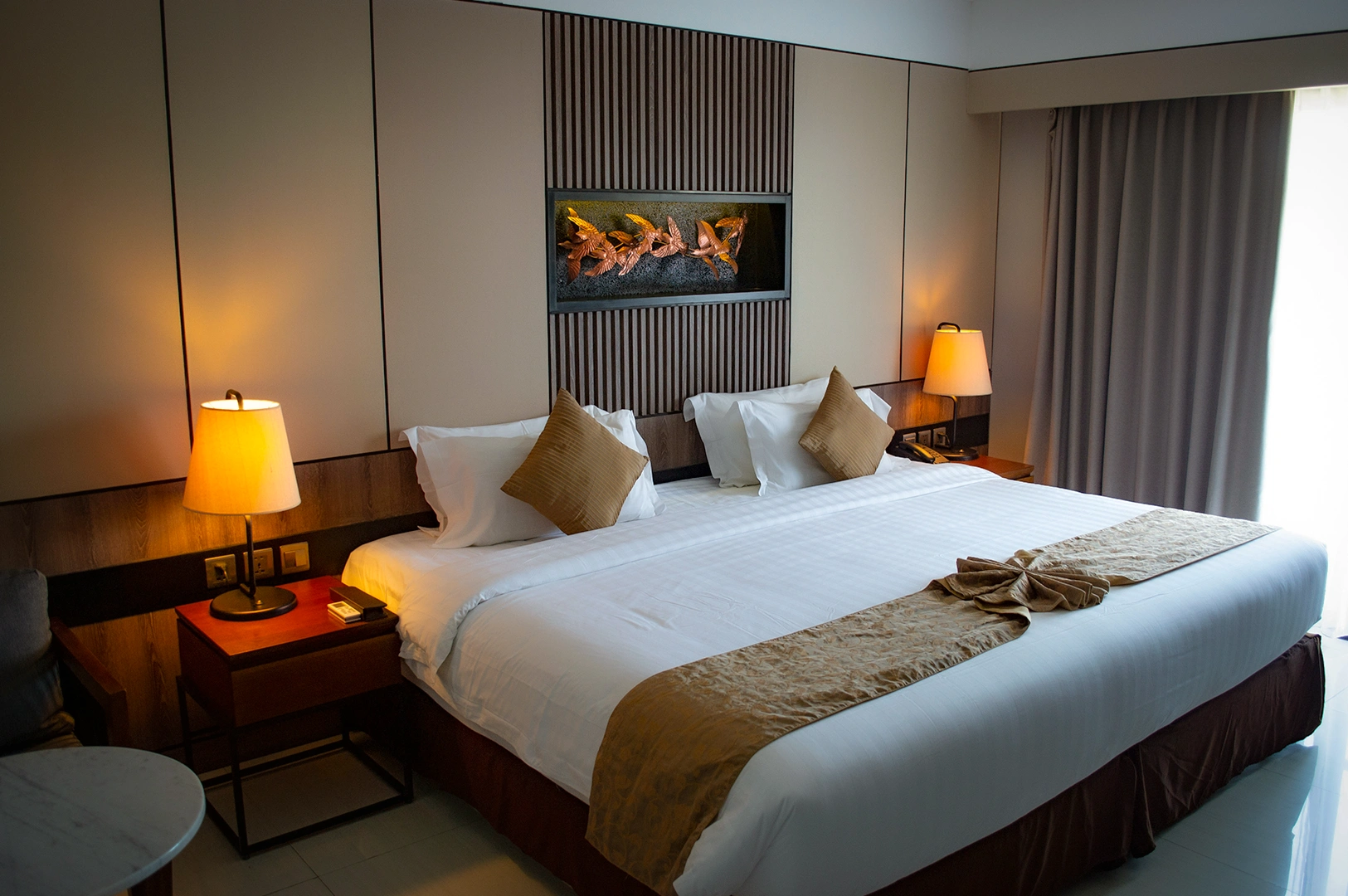 A modern and spacious hotel room interior in Singapore featuring a large king-sized bed with crisp white linens and a gold decorative bed runner. The room is warmly lit by two bedside lamps on dark wood nightstands, highlighting a decorative wall alcove with gold bird sculptures above the headboard.