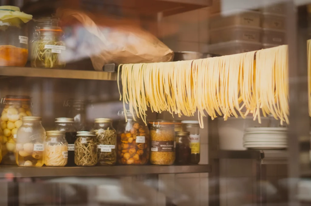 Fresh homemade pasta strands hanging to dry in a rustic kitchen, with glass jars of herbs, spices, and preserved ingredients on wooden shelves, creating an authentic artisanal cooking atmosphere.