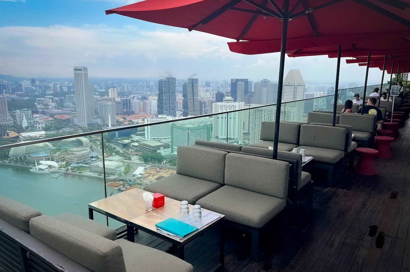 Rooftop bar view of Ce La Vi with red umbrellas and lounge seating overlooking a modern city skyline and waterfront, offering panoramic urban views in a luxury outdoor dining setting.