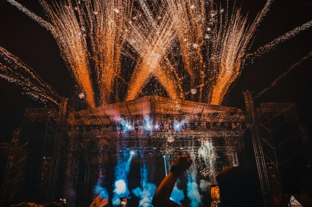 Fireworks exploding above an outdoor concert stage at night with cheering crowd, bright pyrotechnics, and live music festival atmosphere