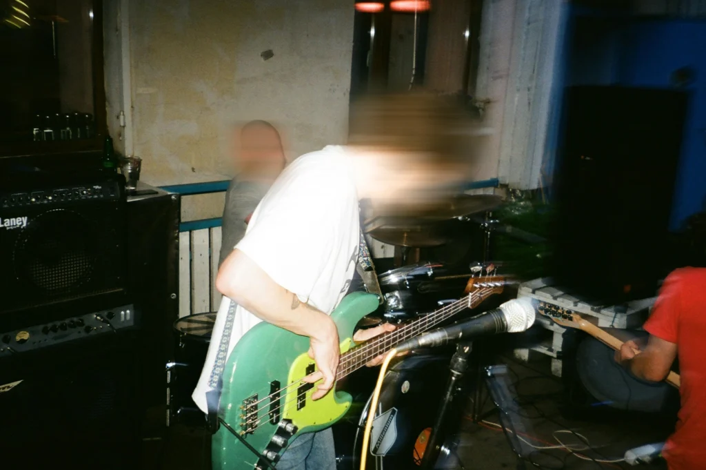 Blurry motion shot of a live band performance in a small venue, featuring a bass guitarist playing a green electric bass with amplifiers, drums, and microphones in a gritty rehearsal space.