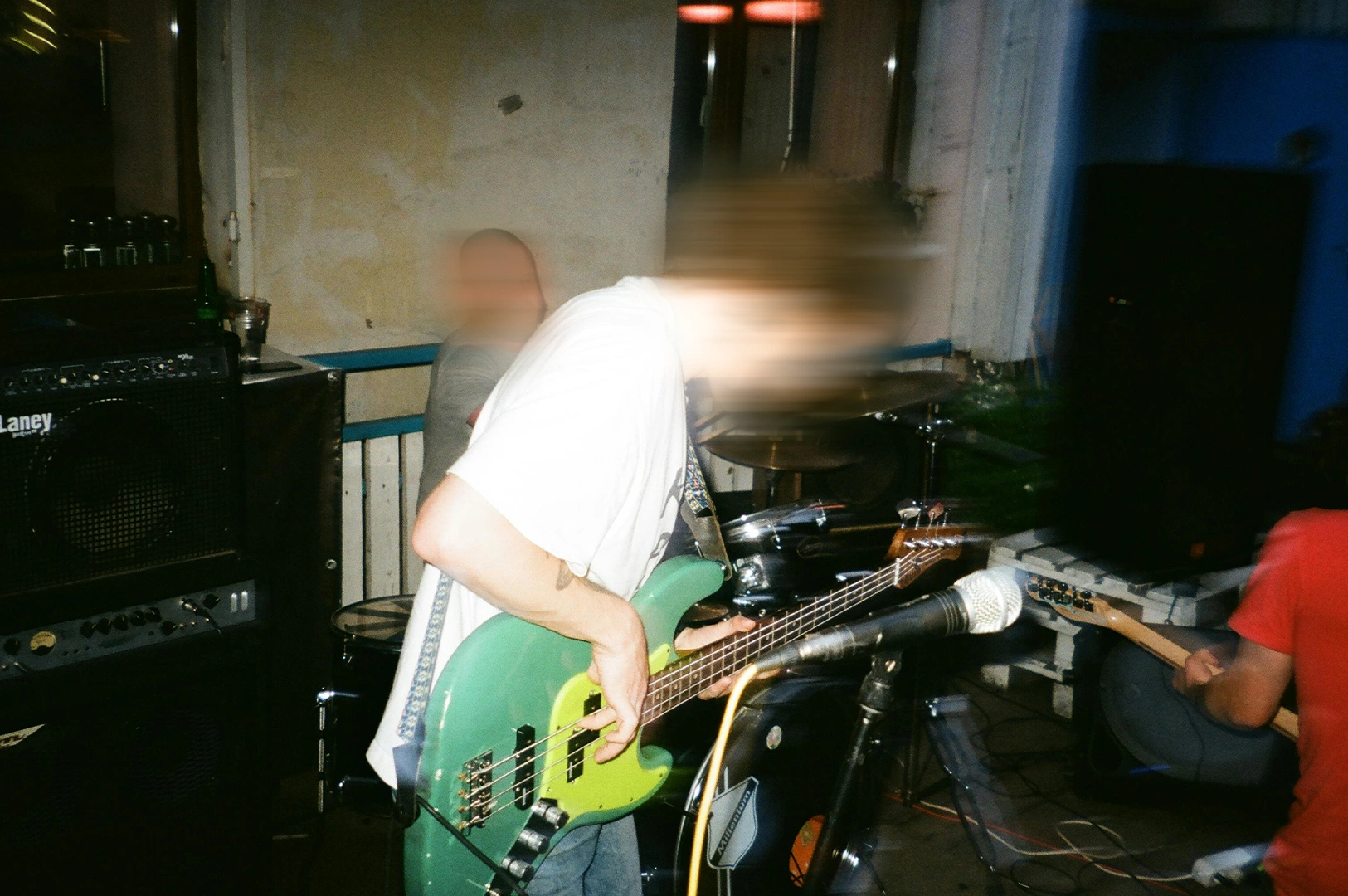Blurry motion shot of a live band performance in a small venue, featuring a bass guitarist playing a green electric bass with amplifiers, drums, and microphones in a gritty rehearsal space.
