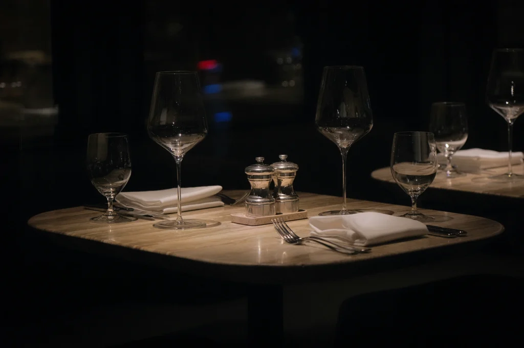 Elegant fine dining table setting with wine glasses, folded white napkins, silverware, and salt and pepper shakers on a dimly lit restaurant table.
