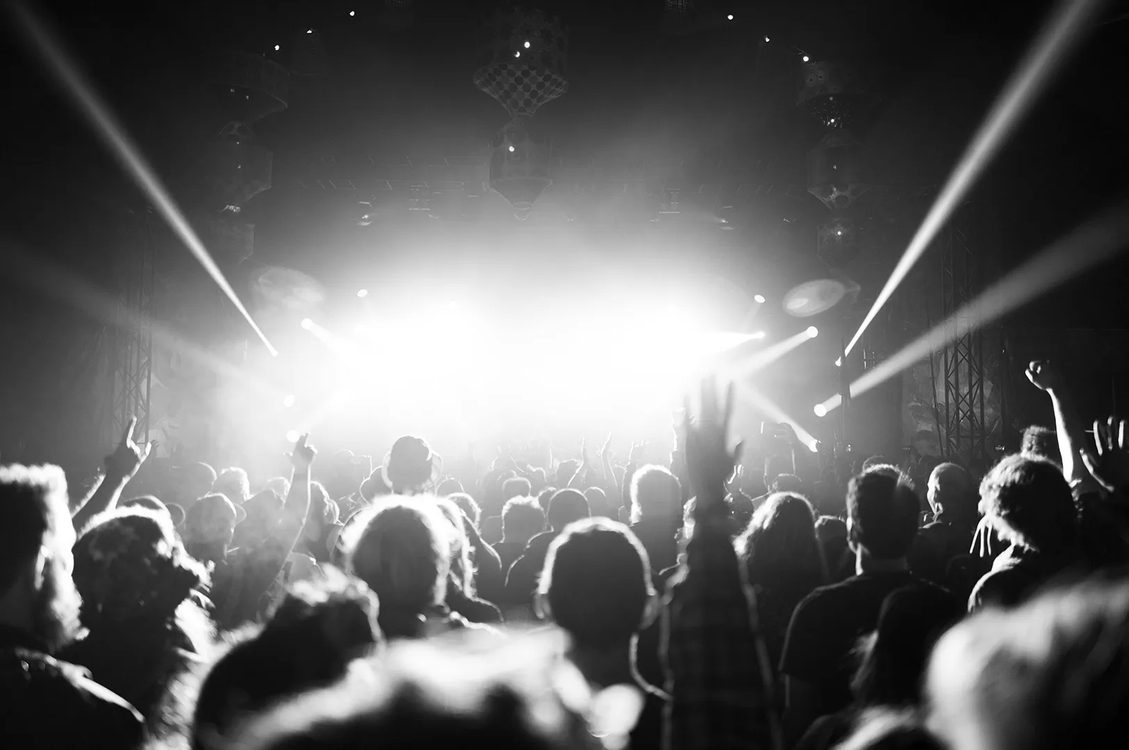 Black and white photo of a live concert crowd with raised hands facing bright stage lights and smoke effects during a music performance