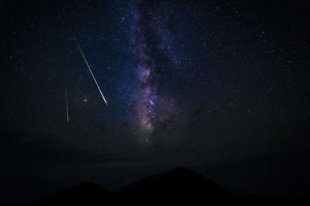 Bright meteor streaks across the Milky Way galaxy above dark mountain silhouettes under a clear star-filled night sky