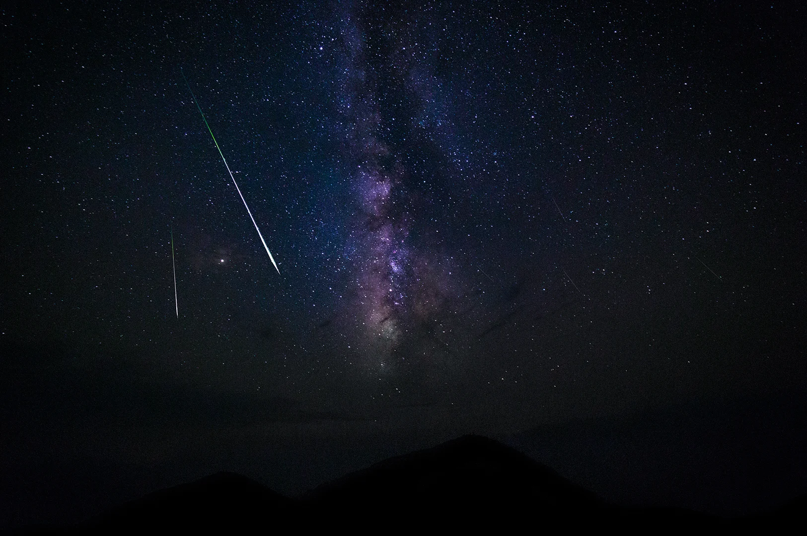Bright meteor streaks across the Milky Way galaxy above dark mountain silhouettes under a clear star-filled night sky